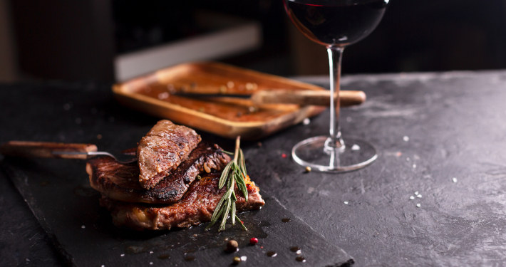 Tranchen von Roastbeef mit Rosmarin auf einer schwarzen Planke mit einem Glas Rotwein im Hintergrund